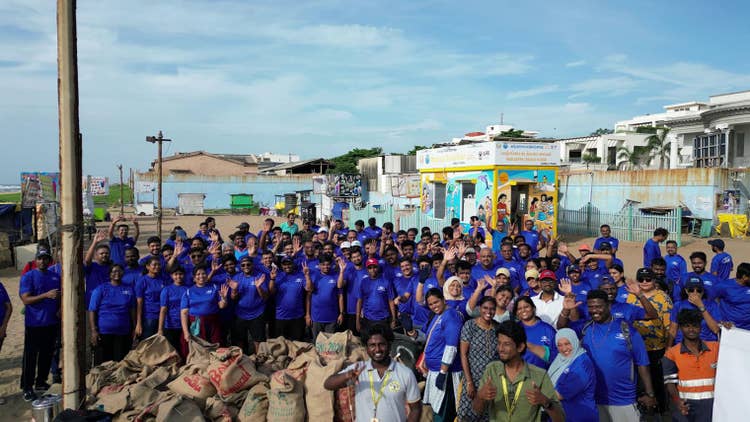 About 80 persons, most wearing blue Ford Volunteer T-shirts, outside on the Manjappai Beach with kiosks in the background.