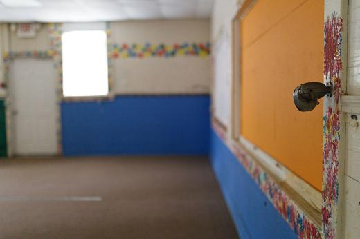Wall-mounted pencil sharpener in a Stanton School classroom that was used to host Head Start program until 2011. Lower half of two walls painted blue. Blue, crimson, yellow and Kelly green splotches frame white and orange areas of a wall, the door frame, a window and decorate the wall in a horizontal line.