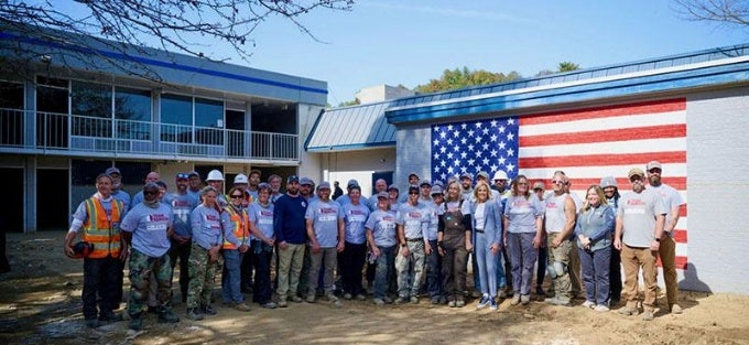 Almost 40 persons in workwear, many wearing Team Rubicon T-shirts, stand outside in front of an L-shaped building, one part painted with an American flag.