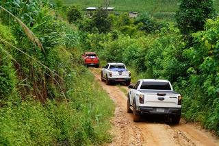 A red truck leads two white trucks on a dirt road with tall vegetation on either side. Farm fields and a couple of buildings are in the background.