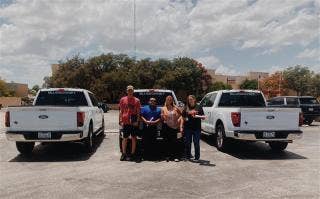 A group of four people stand in front of the middle of three, white Ford trucks with BLUEBONNET on the rear window.