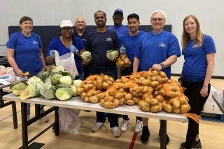 Eight volunteers wearing Ford Volunteer Corps T-shirts, standing behind a table full of cabbage and potatoes.