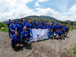 More than 35 people in blue shirts gather for an outdoor group photo while holding a Global Caring Month banner