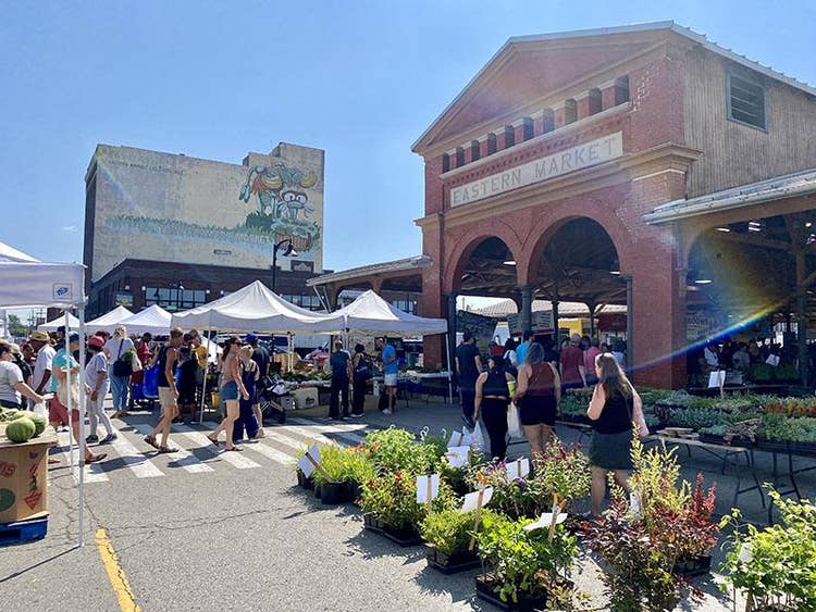 Brick building with double-arched entrance and the words EASTERN MARKET etched in stone, surrounded by pop-up tents and shrubs and tables of plants displayed on the asphalt. A mural of animals shaped from vegetables is on a building in the distance.