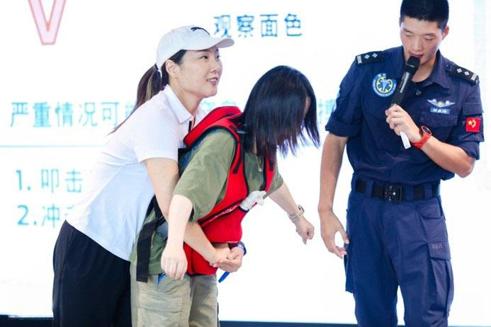 Female has her arms in the Heimlich position around another person while an instructor with a microphone is offside.