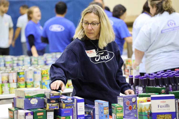 Blonde female wearing navy Ford sweatshirt, making selection from a variety of boxed pasta with several persons wearing blue Ford T-shirts in the background and tables of canned goods.