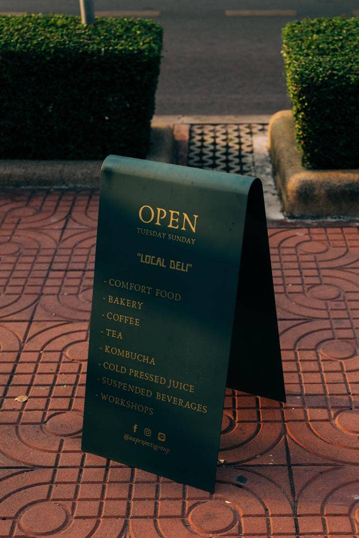 Sitting on dark terra cotta colored sidewalk with intricate geometric design a black sandwich sign with gold text: OPEN TUESDAY SUNDAY “LOCAL DELI’ • COMFORT FOOD • BAKERY • COFFEE • TEA • KOMBUCHA • COLD PRESSED JUICE • SUSPENDED BEVERAGES • WORKSHOPS Icons for facebook, Instagram, social line @naprojectsgroup