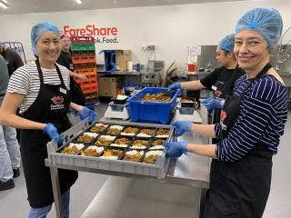 Three people wearing hairnets and FareShare aprons work at a table placing food into containers in the midst of a commercial kitchen, while others work around them.