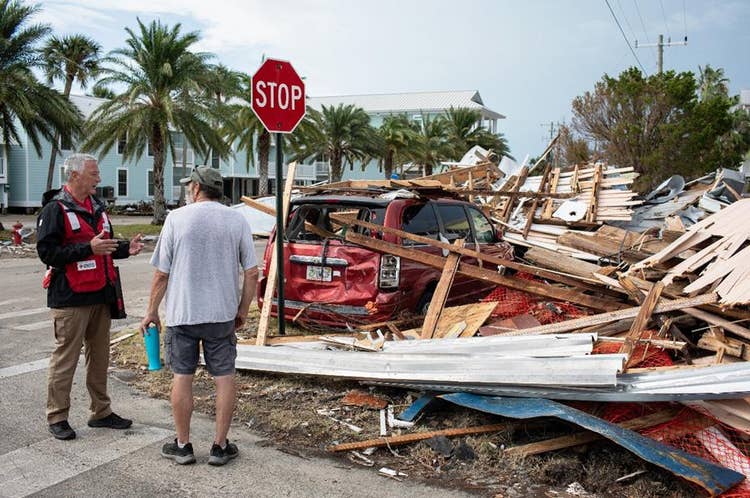 Street corner with hurricane debris and palm trees with a red cross volunteer talking to a resident