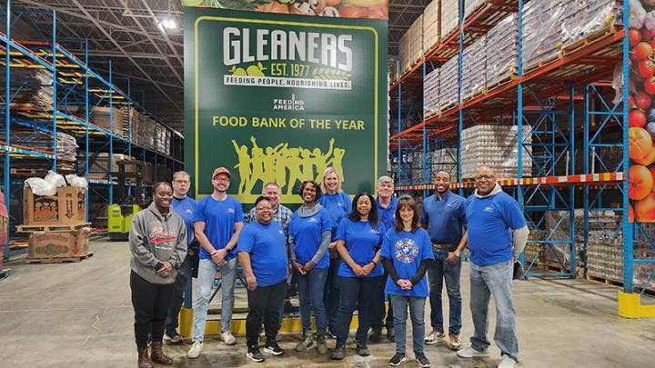 Green hanging banner: GLEANERS EST. 1977 FEEDING PEOPLE. NOURISHING LIVES. Feeding America logo. FOOD BANK OF THE YEAR behind 12 people, most wearing Ford Volunteer Corps T-shirts, in a warehouse