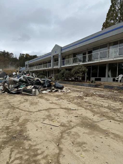 A pile of damaged mattresses, mattress springs, wall air conditioner units, lawn chairs, filled trash bags, etc., in front of a two-story structure.