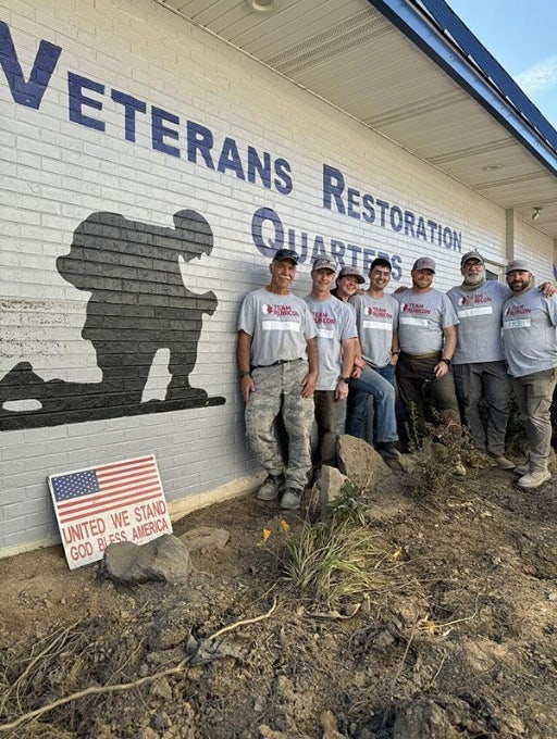 Seven people clad in Team Rubicon T-shirts stand in front of a building with a silhouette of a soldier taking a knee with head bowed and hands clasped together and VETERANS RESTORATION QUARTERS. At their feet is a U.S. flag on a sign with UNITED WE STAND GOD BLESS AMERICA.