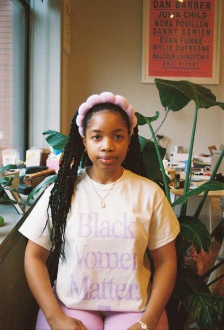 Young female African American, wearing pastel T-shirt with the printing Black Women Matter, seated in front of plants, in front of a desk. On the wall is a poster printed with the names Dan Barber, Julia Child, Nora Pouillon, Danny Bowien, Evan Funke, Wylie Defresne, Malcolm Livingston II, Eric Ripert.