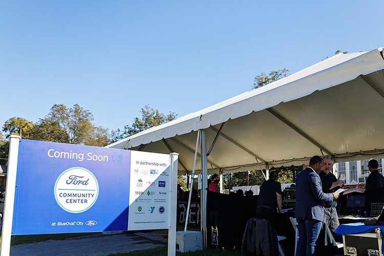 Blue and white sign, Coming Soon Ford COMMUNITY CENTER at BlueOval City with partner logos, sits aside large white tent with a handful of people underneath.
