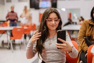 Female student wearing eyeglasses looks into cell phone held in their left hand.