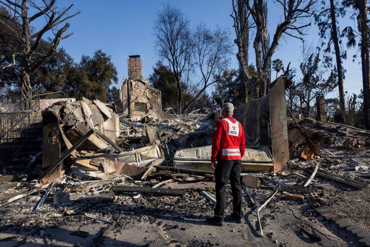 A red cross member stands before a burned house, highlighting the destruction caused by a recent fire emergency.