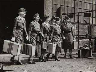 Six Red Cross Women’s Motor Corps workers in uniform, most holding suitcases in their right hands, walking away from a building. June 1942.