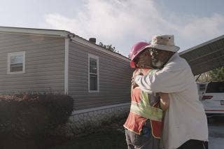 Bearded male wearing eye glasses and fisherman hat hugs person in orange vest and hard hat with carport roof slopped above vehicle beside house.