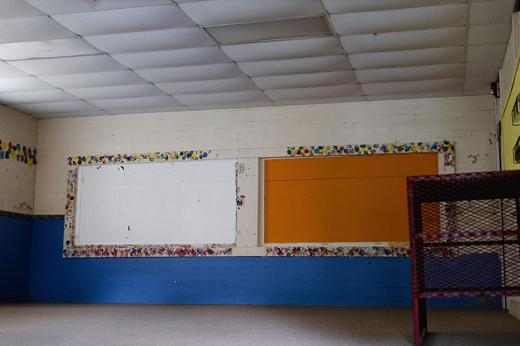 A Stanton School classroom that was used to host Head Start program until 2011. Lower half of two walls painted blue. Blue, crimson, yellow and Kelly green splotches frame white and orange areas of a wall. A rusted red table stands in front of a wall with a yellow section.