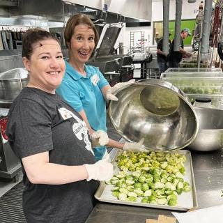 Two hair-netted and gloved Dare to Care Food Bank volunteers preparing romanesco and Brussels sprouts in a commercial kitchen with others in the distant background.