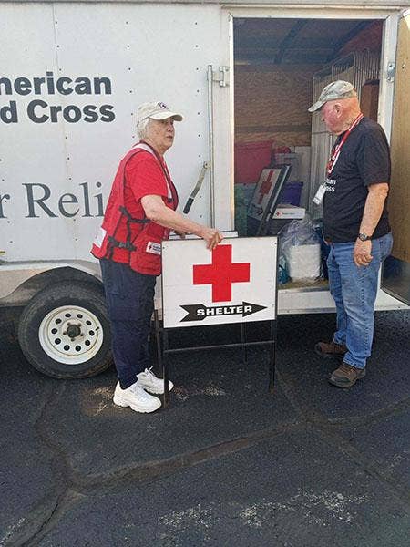 A person in a American Red Cross vest holding a sign with a red cross above a black arrow with white letters SHELTER, in front of an ARC Disaster Relief trailer with an open door. Facing the person is a second person with a lanyard and badge dressed in jeans and a T-shirt.