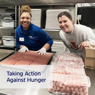 Pair of Dare to Care Food Bank volunteers in a commercial kitchen. African American female, left, wearing gloves, hands on hot dogs in a tray, while second person leans into photo.