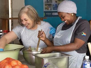 Two persons wearing aprons are stirring and handling food in a room with blue walls. Onions and bowls are on the table.