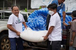 Two people each holding an end of a filled sandbag in front of a truck bed. Each is wearing a white T-shirt with Ford on the front. A blue strip tarp fills the truck bed and two people appear to be talking as one dismounts from the bed.