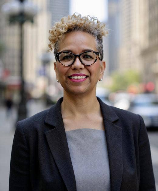African American female wearing a navy blazer over a light gray top, glasses, hair tightly cropped on the sides with curls atop and gently falling face forward on her right side.