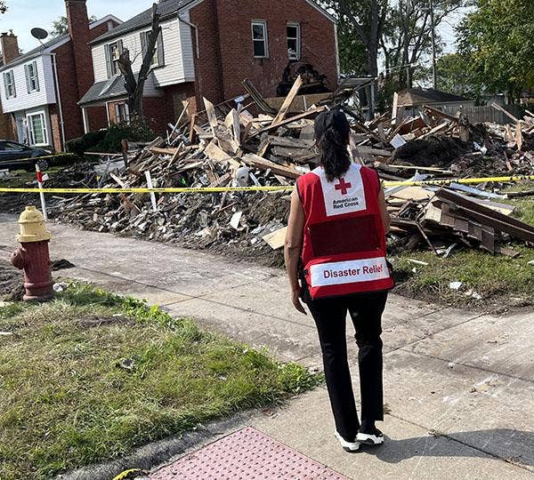Person wearing a red and white American Red Cross Disaster Relief vest with back to camera walks up a sidewalk toward a raized building. Yellow caution tape surrounds the debris area to the right of two houses.