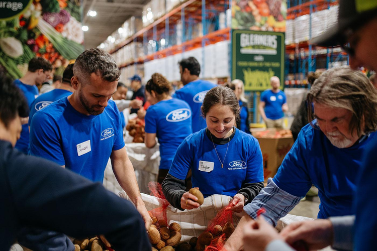 A trio of people in blue Ford Volunteer Corps T-shirts sort potatoes into red, mesh produce bags. Other Volunteer Corps members are visible in the background, in a Gleaner’s warehouse, also sorting potatoes.