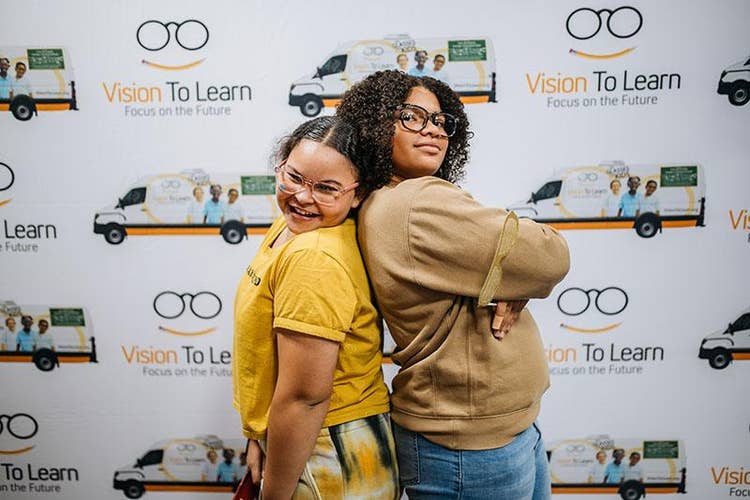 Two female youths, wearing eye glasses, standing back-to-back in front of a Vision To Learn Focus on the Future backdrop at Vision Party.