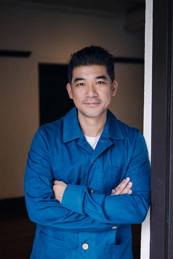 Sakson Rouypirom leans against a doorframe on his left site and has a friendly smile while wearing a blue chambray work shirt as the collar of a white T-shirt peeks ouf from underneath.