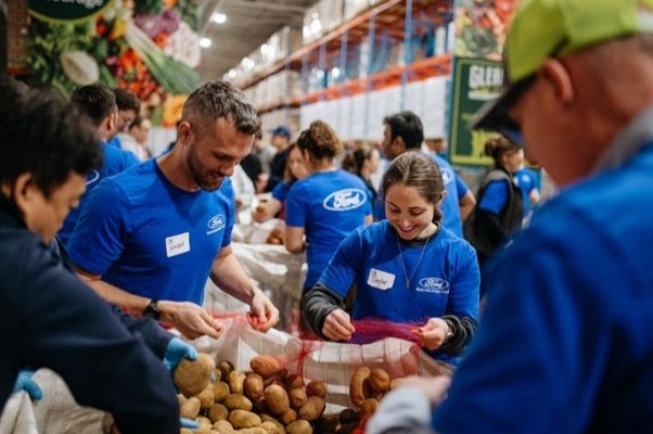A male and a female wearing Ford Volunteer Corps T-shirts while sorting potatoes into mesh bags, in a warehouse with others in the foreground and background also working