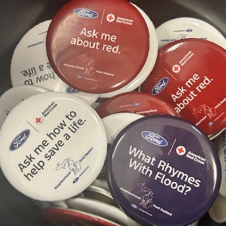 Red, white and blue pin on buttons in a container with Ford blue ovals and American Red Cross logos with Ask me about red. And Ask me how to help save a life. and what Rhymes With Flood.