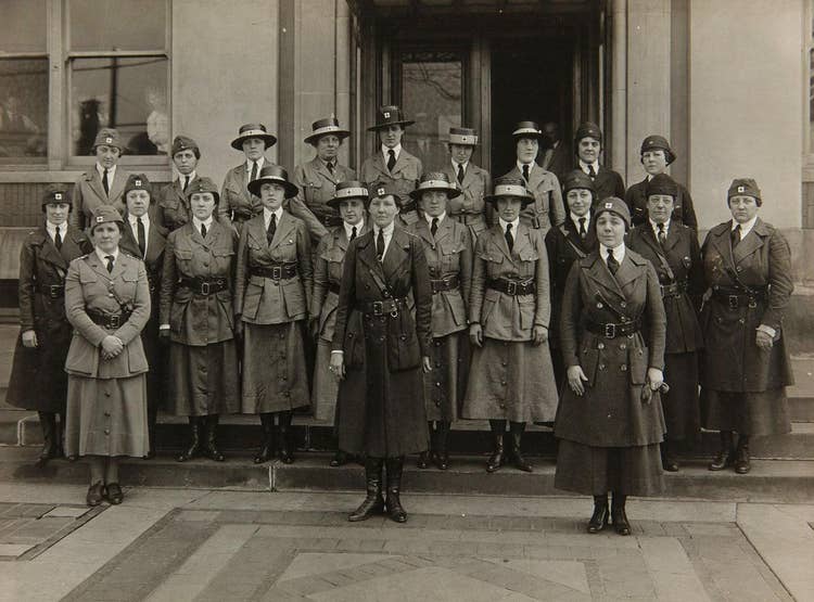 Twenty-two Red Cross workers on the Ford Highland Park Plant steps in 1918, while people look out the windows.