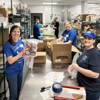 Trio of Dare to Care Food Bank volunteers organizing food donations with another trio in the background prepping food, all in commercial kitchen.