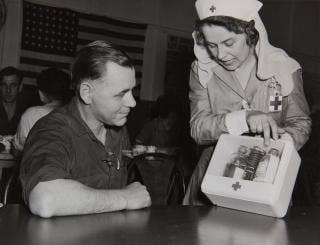 25,000th Blood Donor, seated male in work shirt with rolled up right sleeve in the Ford Rouge Plant Pressed Steel Building, May 1943, with a Red Cross nurse showing him a kit.