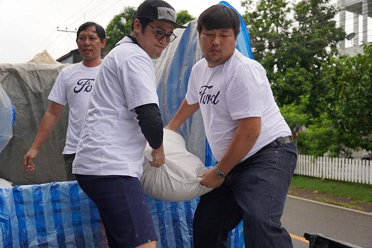 Two males each holding two corners of a sandbag. A third person stands behind them. They all wear white T-shirts with Ford in script on the front of the shirts as they stand in a truck bed.