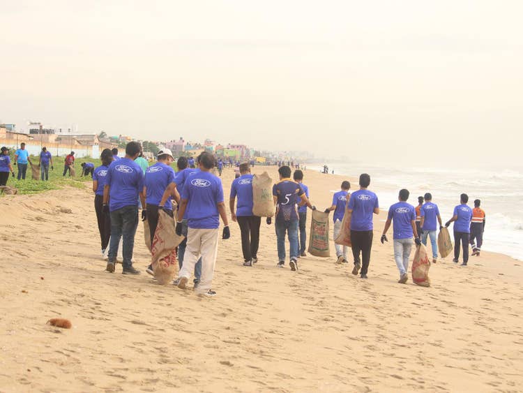 More than a dozen people wearing blue Ford Volunteer T-shirts walk the beach with trash bags while others are on the grass picking up trash.