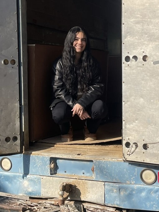 Smiling female with long, dark hair wearing dark clothing and tan boots, squatting in cargo container, framed by the open doors.