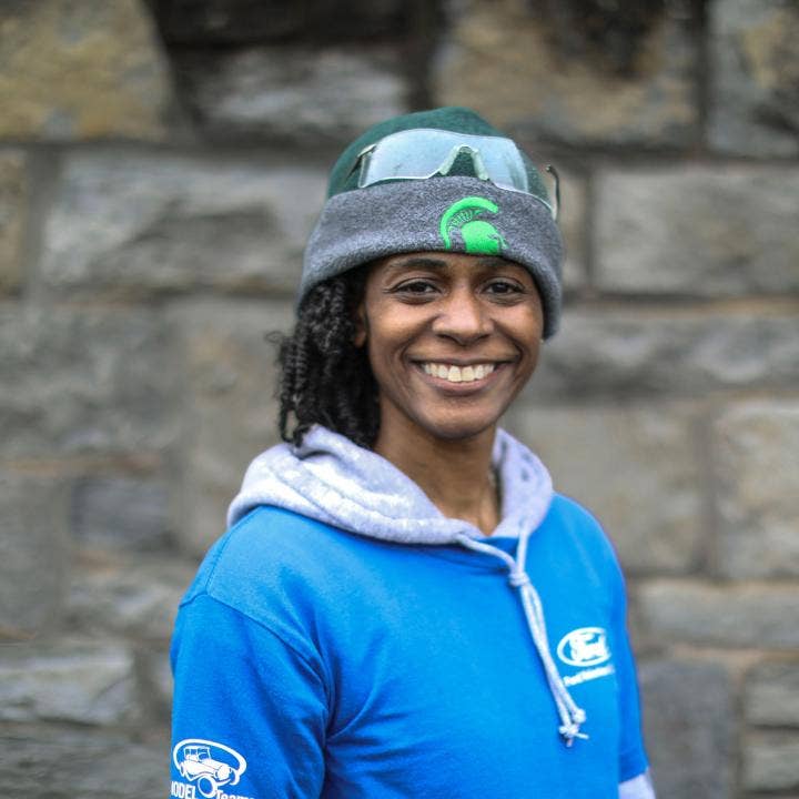African American female wearing Michigan State University green and gray knit cap with Spartan logo, aqua and gray Ford Volunteer logo hoodie, standing in front of a brick wall.