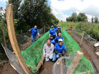 Nine persons, some in blue Ford Philanthropy T-shirts, all in a field bordered by trees. Green netting covers two rows of plants with all but one of the people between the rows. Chicken wire fencing on either side of the protected rows.
