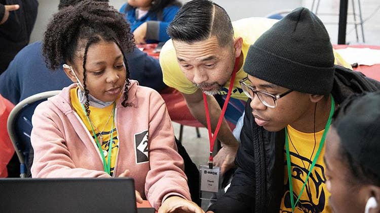 Asian male bends down between two African American students while a third looks on as all focus on a laptop.