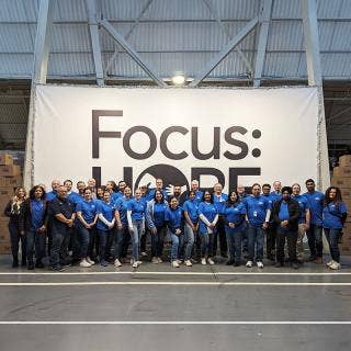 More than 30 people in blue Ford Volunteer T-shirts and mostly jeans in two rows in front of a 24 ft. Focus: HOPE banner.
