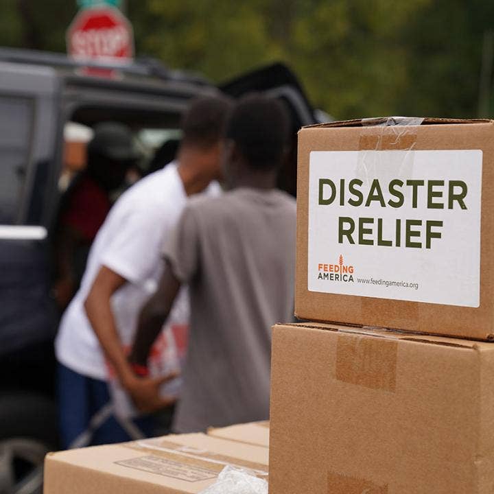 Two cardboard boxes stacked atop each other with the top box labeled DISASTER RELIEF FEEDING AMERICA www. Feedingamerica.org with more boxes and people working in the background.