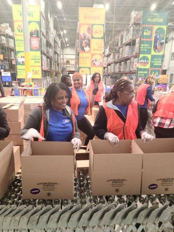 Several people wearing blue Ford Volunteer Corps T-shirts and moving cardboard Forgotten Harvest logo boxes on a conveyor belt in a warehouse