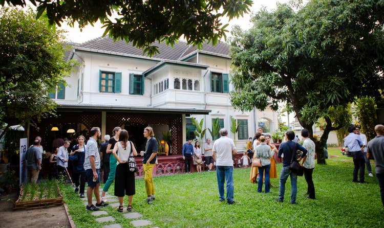 Na Café in daylight with several adults holding beverages and handbags while gathered in small, standing groupings outside a white building with aqua-green shutters. Stones lead to a maroon lattice porch while a maroon boarder frames the grass as a child struts past.