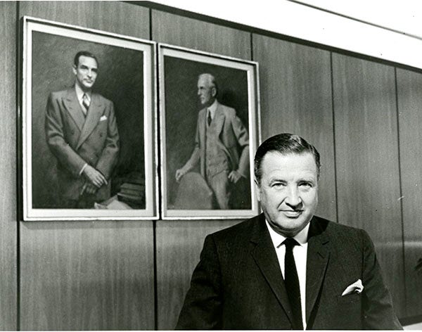 Henry Ford II in front of portraits of his father, Edsel Ford, and his grandfather Henry Ford, in a wood paneled room.