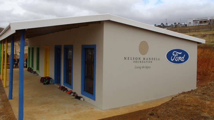 Side view of the school building with children’s shoes lined up against the building on the porch and the end emblazoned with the Nelson Mandela Foundation and Ford logs.